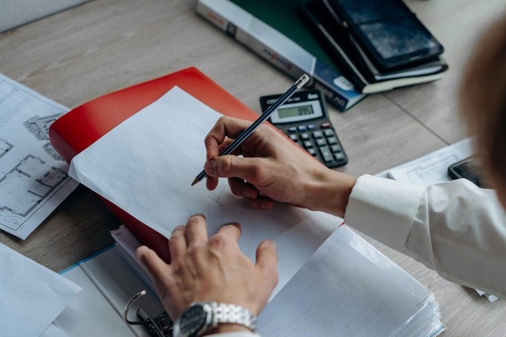 Close-up of hands writing on paper at a desk with a calculator, notebook, and drawing plans.
