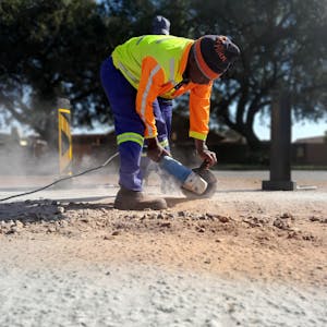 A construction worker in safety gear operates a power tool on a road surface.