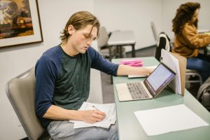College student writing notes and using a laptop at a classroom desk for exam preparation.