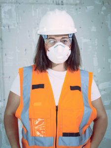 Female engineer in safety gear with hard hat and N95 mask indoors.